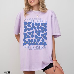 May include: A lavender-colored t-shirt with a graphic design of blue flowers and the text "Where the Spirit of the Lord is There is Freedom 2 Corinthians 5:17".