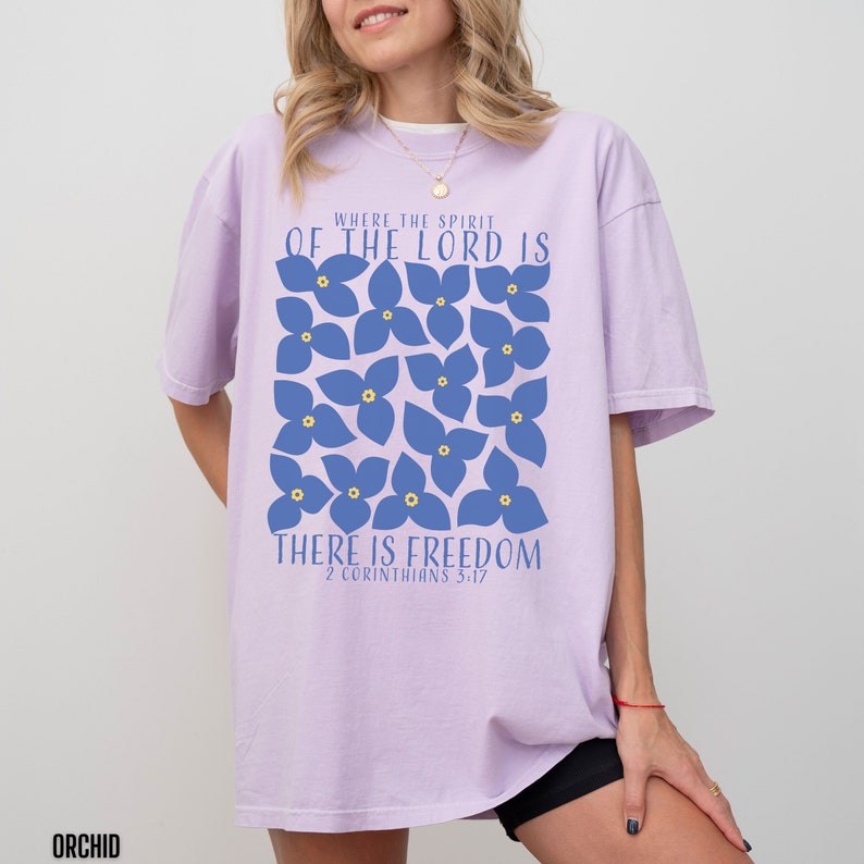 May include: A lavender-colored t-shirt with a graphic design of blue flowers and the text "Where the Spirit of the Lord is There is Freedom 2 Corinthians 5:17".