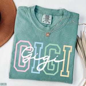 May include: A light green t-shirt with the word "Gigi" written in white script with rainbow colored outlines. The shirt has a tag that says "Comfort Colors" and "S - P - CH".