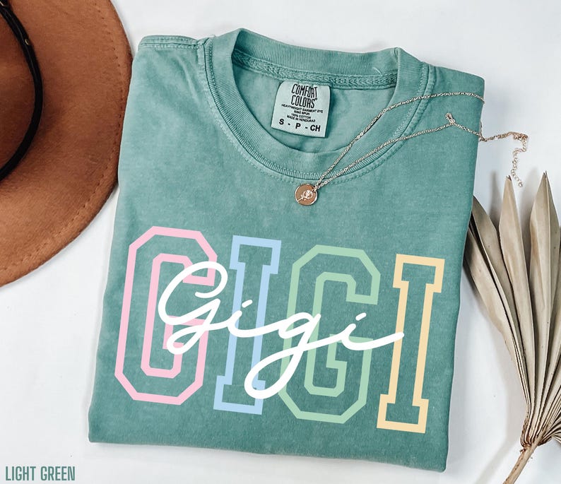 May include: A light green t-shirt with the word "Gigi" written in white script with rainbow colored outlines. The shirt has a tag that says "Comfort Colors" and "S - P - CH".