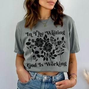 Op de afbeelding: Een grijs T-shirt met een zwart bloemenontwerp en de tekst "In The Waiting God Is Working".