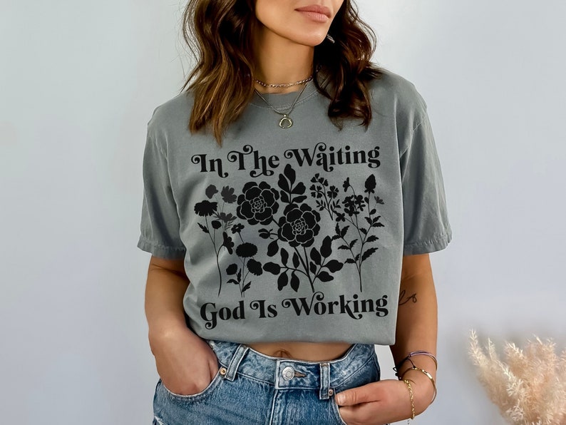 Op de afbeelding: Een grijs T-shirt met een zwart bloemenontwerp en de tekst "In The Waiting God Is Working".