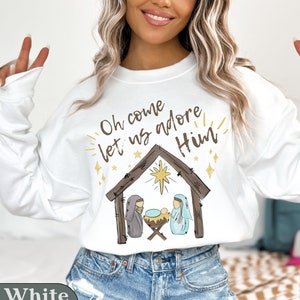 Puede incluir: Sudadera blanca con un gr&aacute;fico de la natividad de Navidad y el texto "Oh come let us adore Him".