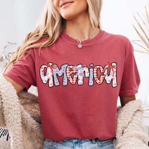 May include: Red t-shirt with a graphic of the word "America" in blue and white with a red, white, and blue checkered pattern. The graphic has a festive, patriotic theme with fireworks.