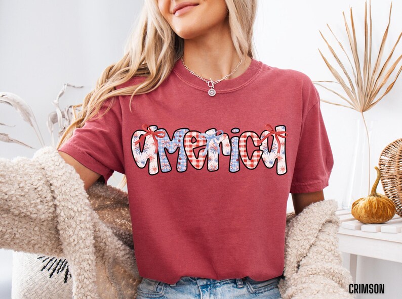 May include: Red t-shirt with a graphic of the word "America" in blue and white with a red, white, and blue checkered pattern. The graphic has a festive, patriotic theme with fireworks.