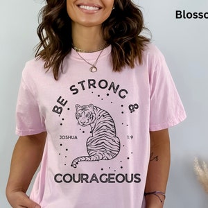 May include: Pink t-shirt with a black and white tiger graphic and the text "Be Strong & Courageous" with the words "Joshua 1:9" underneath the tiger.