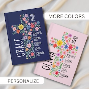 May include: Two journals with floral cross designs. One is navy blue with "GRACE" and the words "YOU ARE... BEAUTIFUL, STRONG, CHOSEN, LOVED, FORGIVEN." The other is pink with "OLIVIA" and similar text. The image also includes the words "MORE COLORS" and "PERSONALIZE."