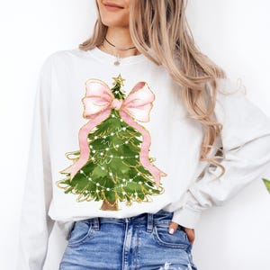 K&ouml;nnte beinhalten: Wei&szlig;es Langarm-Sweatshirt mit einem Aquarell-Weihnachtsbaum-Design. Der Baum ist gr&uuml;n mit goldenen Akzenten, einer rosa Schleife und Lichterketten. Das Wort "WHITE" ist unten links im Bild aufgedruckt.