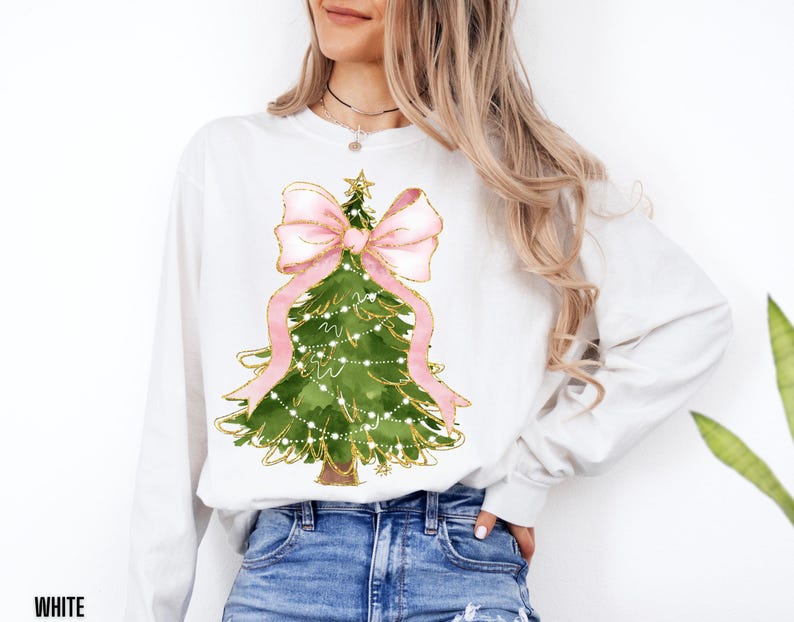 K&ouml;nnte beinhalten: Wei&szlig;es Langarm-Sweatshirt mit einem Aquarell-Weihnachtsbaum-Design. Der Baum ist gr&uuml;n mit goldenen Akzenten, einer rosa Schleife und Lichterketten. Das Wort "WHITE" ist unten links im Bild aufgedruckt.