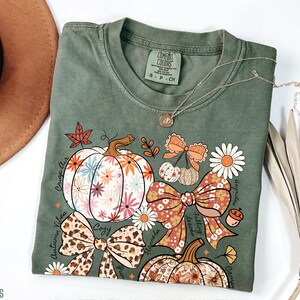 Puede incluir: Camiseta verde musgo con un dise&ntilde;o de tem&aacute;tica oto&ntilde;al. El dise&ntilde;o incluye calabazas florales, lazos y las palabras "Crisp Air", "Autumn Vibes" y "Cozy". Un collar plateado con un colgante est&aacute; sobre la camiseta.