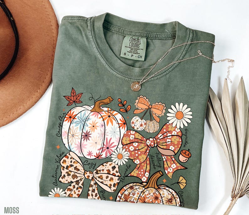 Puede incluir: Camiseta verde musgo con un dise&ntilde;o de tem&aacute;tica oto&ntilde;al. El dise&ntilde;o incluye calabazas florales, lazos y las palabras "Crisp Air", "Autumn Vibes" y "Cozy". Un collar plateado con un colgante est&aacute; sobre la camiseta.
