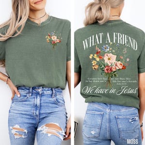 K&ouml;nnte beinhalten: Olivgr&uuml;nes T-Shirt mit floralem Bouquet-Design auf Vorder- und R&uuml;ckseite. Auf der R&uuml;ckseite des Shirts steht der Text "WHAT A FRIEND" &uuml;ber dem floralen Design, mit zus&auml;tzlichem Text darunter: "We have in Jesus".