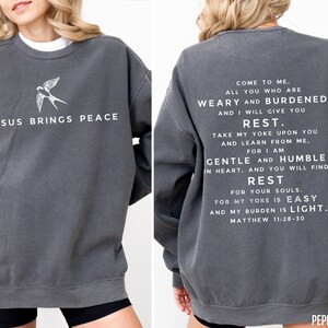 May include: A dark gray sweatshirt with a white bird graphic on the front and a white text quote from the Bible on the back. The quote is from Matthew 11:28-30 and reads "Come to me, all you who are weary and burdened, and I will give you rest. Take my yoke upon you and learn from me, for I am gentle and humble in heart, and you will find rest for your souls. For my yoke is easy and my burden is light."