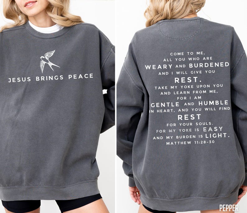 May include: A dark gray sweatshirt with a white bird graphic on the front and a white text quote from the Bible on the back. The quote is from Matthew 11:28-30 and reads "Come to me, all you who are weary and burdened, and I will give you rest. Take my yoke upon you and learn from me, for I am gentle and humble in heart, and you will find rest for your souls. For my yoke is easy and my burden is light."