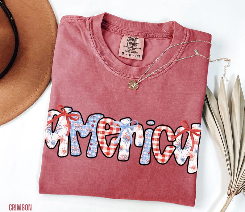 May include: A red t-shirt with the word "america" written in a colorful patchwork style. The letters are decorated with red, white, and blue gingham, fireworks, and bows. The shirt is made by Comfort Colors.