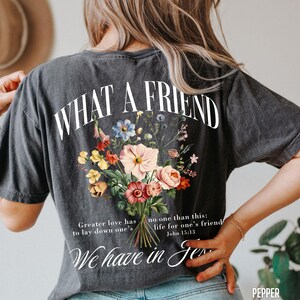 K&ouml;nnte beinhalten: Dunkelgraues T-Shirt mit dem Text "WHAT A FRIEND" &uuml;ber einem Blumenstrau&szlig;-Design. Der Strau&szlig; zeigt rosa, gelbe und blaue Blumen. Darunter stehen die Worte "Greater love has to lay down one's" und "We have in Je".