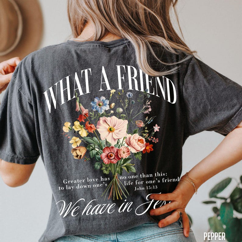 K&ouml;nnte beinhalten: Dunkelgraues T-Shirt mit dem Text "WHAT A FRIEND" &uuml;ber einem Blumenstrau&szlig;-Design. Der Strau&szlig; zeigt rosa, gelbe und blaue Blumen. Darunter stehen die Worte "Greater love has to lay down one's" und "We have in Je".