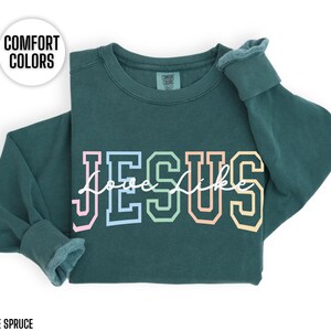 May include: A dark green sweatshirt with the word "JESUS" in rainbow coloured letters and the words "Love Like" written in white script underneath. The sweatshirt is made of a soft, comfortable material.