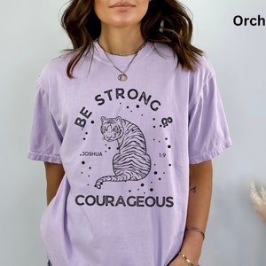 May include: A lavender colored t-shirt with a black graphic of a tiger and the text "Be Strong & Courageous" with the words "Joshua 1:9" underneath the tiger.