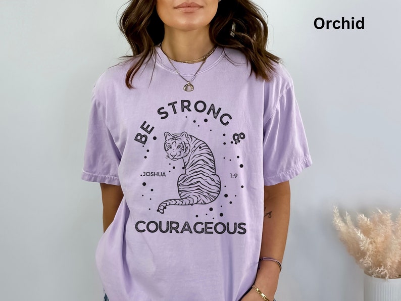 May include: A lavender colored t-shirt with a black graphic of a tiger and the text "Be Strong & Courageous" with the words "Joshua 1:9" underneath the tiger.