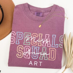 May include: Berry-coloured t-shirt with the words "Specials Miss Squad Anna Art" in a colourful, collegiate-style font. A gold necklace is draped across the shirt. A brown hat and dried palm leaves are in the background.