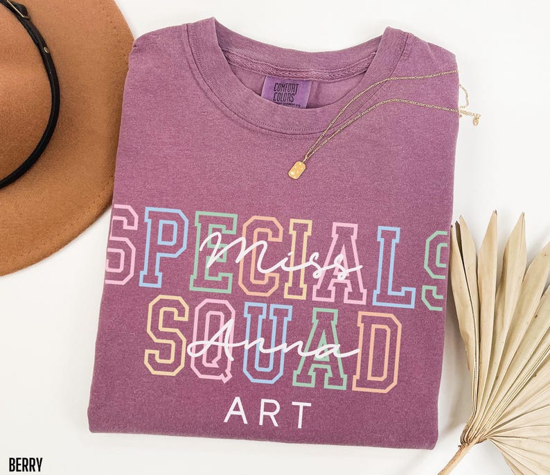 May include: Berry-coloured t-shirt with the words "Specials Miss Squad Anna Art" in a colourful, collegiate-style font. A gold necklace is draped across the shirt. A brown hat and dried palm leaves are in the background.
