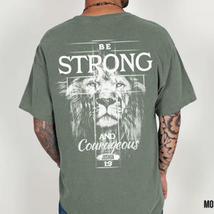 May include: Moss green t-shirt with a lion graphic and the words "BE STRONG AND Courageous" in white. The text "Joshua 1:9" is below the text. The shirt is a short-sleeved crew neck.