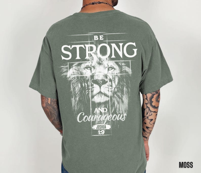 May include: Moss green t-shirt with a lion graphic and the words "BE STRONG AND Courageous" in white. The text "Joshua 1:9" is below the text. The shirt is a short-sleeved crew neck.