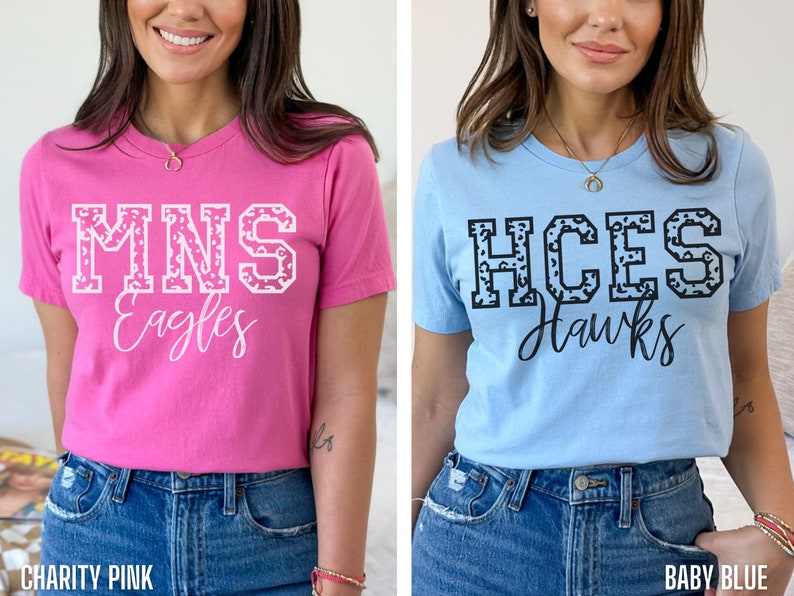 May include: Two t-shirts, one pink and one light blue. The pink shirt has "MNS Eagles" in white block letters with a leopard print pattern. The light blue shirt has "HCES Hawks" in black block letters with a leopard print pattern.
