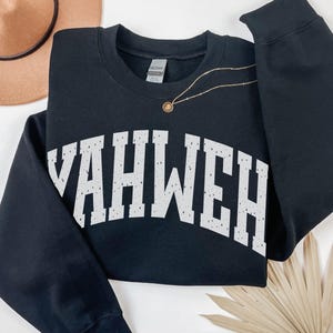 May include: Black sweatshirt with the word "YAHWEH" in distressed white lettering across the chest. The sweatshirt has long sleeves and a crew neck. A gold necklace with a pendant is draped on the sweatshirt. The word "BLACK" is in the bottom right corner.