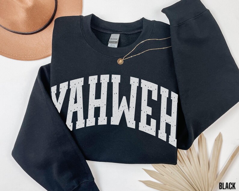 May include: Black sweatshirt with the word "YAHWEH" in distressed white lettering across the chest. The sweatshirt has long sleeves and a crew neck. A gold necklace with a pendant is draped on the sweatshirt. The word "BLACK" is in the bottom right corner.