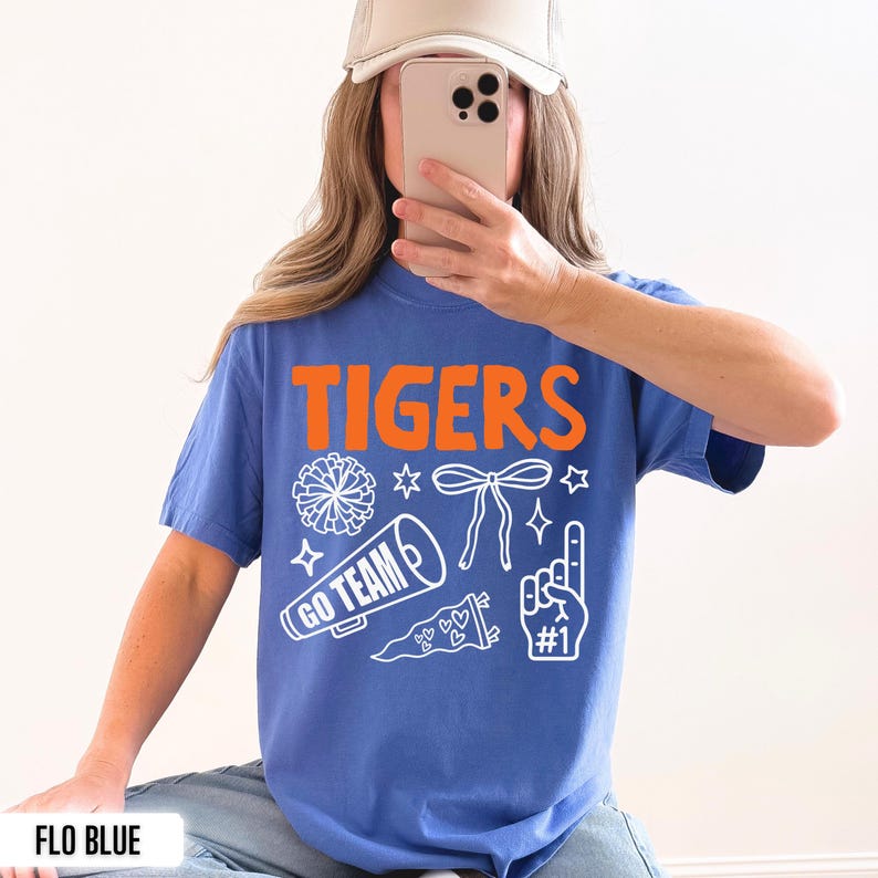 Puede incluir: Una camiseta azul de manga corta con el texto naranja TIGERS e ilustraciones gr&aacute;ficas blancas de un pomp&oacute;n, un lazo, un meg&aacute;fono con el texto GO TEAM, un bander&iacute;n con corazones y un dedo de espuma con el texto #1.