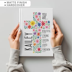May include: A white hardcover journal with a matte finish. The cover features a floral cross design with colorful flowers. The name "Kailey" is printed vertically on the left. Text includes "You Are... Beautiful, Strong, Chosen, Loved, Forgiven" with Bible verses.