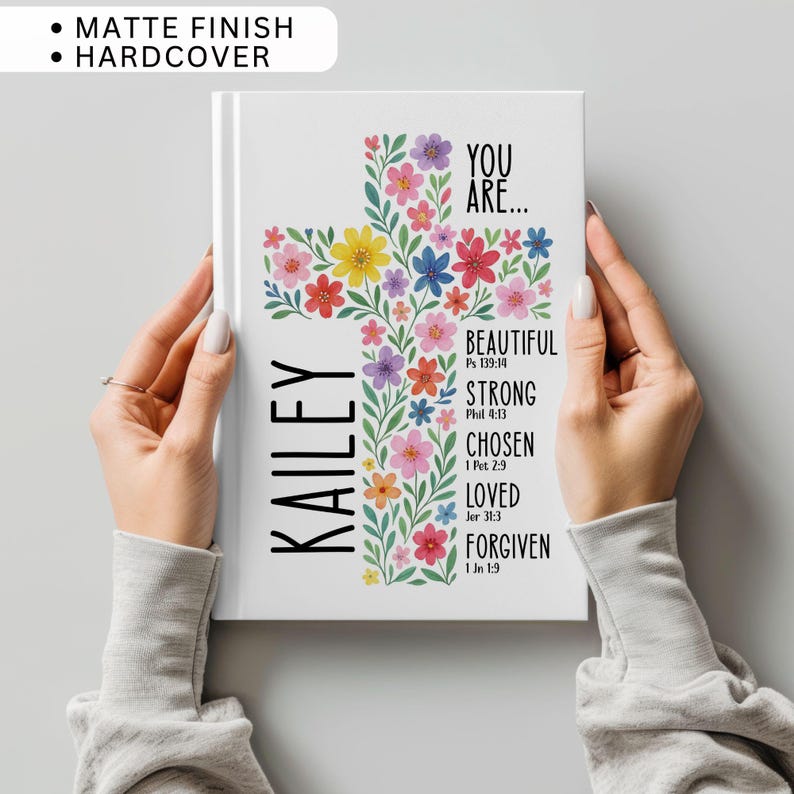 May include: A white hardcover journal with a matte finish. The cover features a floral cross design with colorful flowers. The name "Kailey" is printed vertically on the left. Text includes "You Are... Beautiful, Strong, Chosen, Loved, Forgiven" with Bible verses.