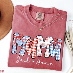 May include: A red Comfort Colors t-shirt with the word "MAMA" in a patriotic design. The letters are decorated with gingham, fireworks, and bows. The names "Jack" and "Anna" are written below the word "MAMA". A gold necklace is draped across the shirt.