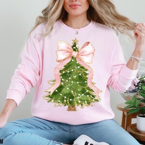 K&ouml;nnte beinhalten: Hellrosa Sweatshirt mit einem Weihnachtsbaum-Design. Der Baum ist gr&uuml;n mit goldenen Akzenten, einer rosa Schleife und Lichterketten. Das Wort "BLOSSOM" ist unten links aufgedruckt.