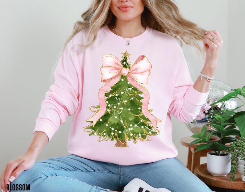 K&ouml;nnte beinhalten: Hellrosa Sweatshirt mit einem Weihnachtsbaum-Design. Der Baum ist gr&uuml;n mit goldenen Akzenten, einer rosa Schleife und Lichterketten. Das Wort "BLOSSOM" ist unten links aufgedruckt.
