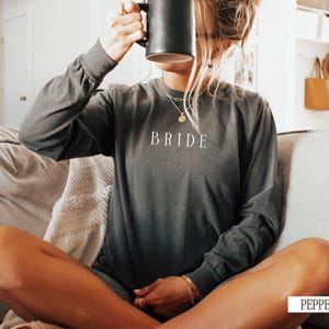 May include: A woman wearing a dark gray long-sleeved shirt with the word "BRIDE" printed on the front. She is sitting on a couch and holding a black mug.