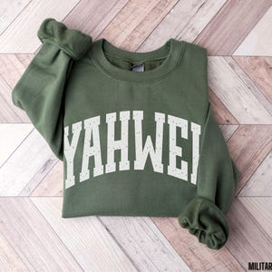 May include: A military green sweatshirt with the word "YAHWEH" printed in white across the chest. The sweatshirt is folded on a light brown and white wooden surface. The sleeves are cuffed.