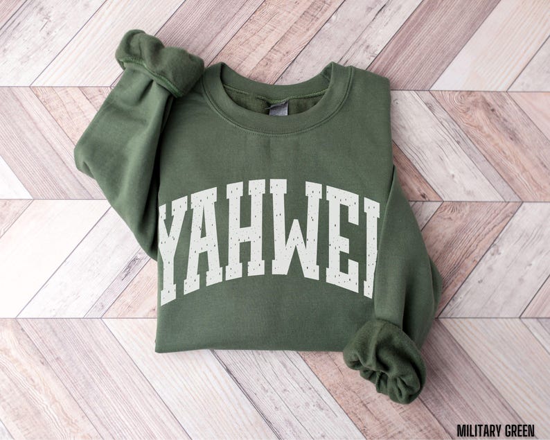 May include: A military green sweatshirt with the word "YAHWEH" printed in white across the chest. The sweatshirt is folded on a light brown and white wooden surface. The sleeves are cuffed.