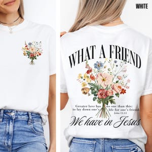 K&ouml;nnte beinhalten: Wei&szlig;es T-Shirt mit einem Blumengesteck-Design auf der Vorder- und R&uuml;ckseite. Auf der R&uuml;ckseite des Shirts steht der Text "WHAT A FRIEND" &uuml;ber einem Blumenarrangement und die Worte "We have in Jesus".