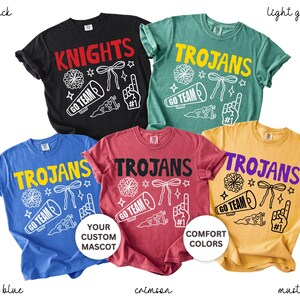 Puede incluir: Cinco camisetas de manga corta en negro, verde claro, azul, carmes&iacute; y mostaza. Cada camiseta presenta un gr&aacute;fico serigrafiado con un meg&aacute;fono, pomp&oacute;n, lazo, bander&iacute;n y dedo de espuma, junto con el texto KNIGHTS o TROJANS y GO TEAM.