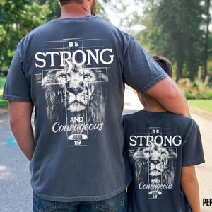 May include: Two gray t-shirts with the text "BE STRONG" above a lion's face and the words "AND Courageous" below. The shirts are worn by a parent and child, suggesting a matching set.