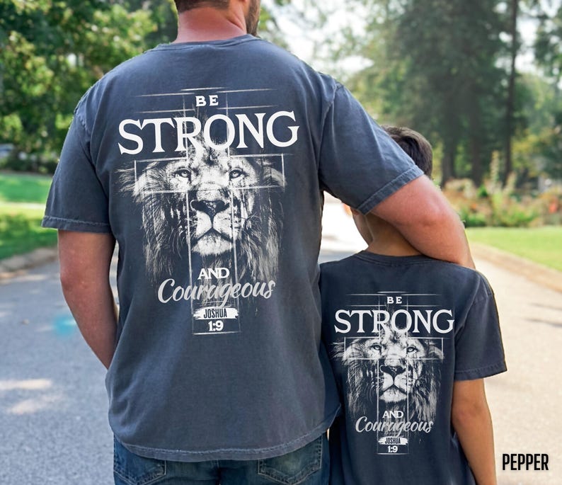 May include: Two gray t-shirts with the text "BE STRONG" above a lion's face and the words "AND Courageous" below. The shirts are worn by a parent and child, suggesting a matching set.