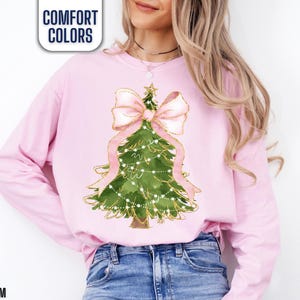 K&ouml;nnte beinhalten: Ein hellrosa Langarm-Sweatshirt mit einem Weihnachtsbaum-Design. Der gr&uuml;ne Baum ist mit wei&szlig;en Lichtern, einer rosa Schleife und einem goldenen Stern verziert. Der Text "COMFORT COLORS" befindet sich in einer wei&szlig;en Box und das Wort "BLOSSOM" steht unten.