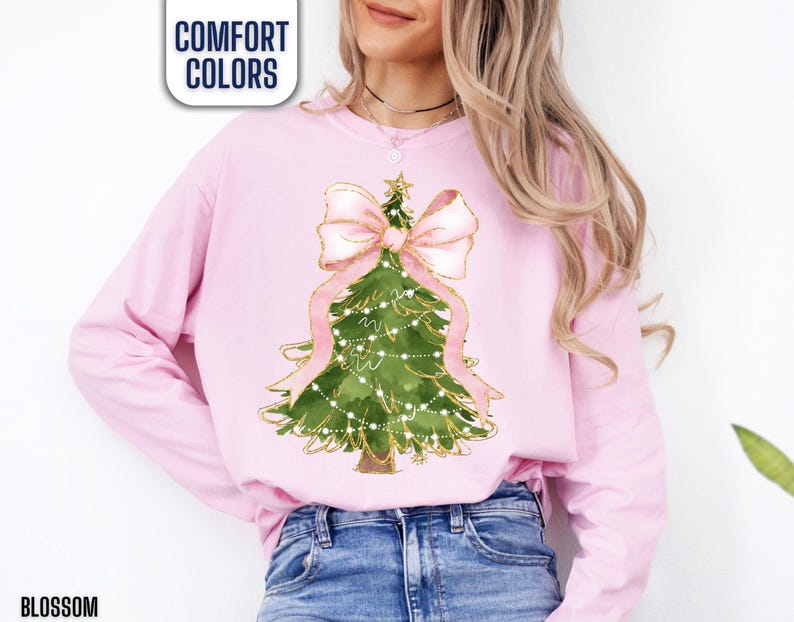 K&ouml;nnte beinhalten: Ein hellrosa Langarm-Sweatshirt mit einem Weihnachtsbaum-Design. Der gr&uuml;ne Baum ist mit wei&szlig;en Lichtern, einer rosa Schleife und einem goldenen Stern verziert. Der Text "COMFORT COLORS" befindet sich in einer wei&szlig;en Box und das Wort "BLOSSOM" steht unten.