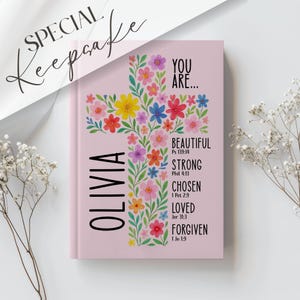 May include: A pink journal with a floral cross design. The cross is filled with colorful flowers. The journal has the name "OLIVIA" vertically on the left side and the words "YOU ARE... BEAUTIFUL, STRONG, CHOSEN, LOVED, FORGIVEN" on the right.