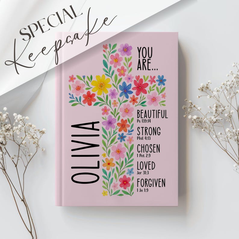 May include: A pink journal with a floral cross design. The cross is filled with colorful flowers. The journal has the name "OLIVIA" vertically on the left side and the words "YOU ARE... BEAUTIFUL, STRONG, CHOSEN, LOVED, FORGIVEN" on the right.