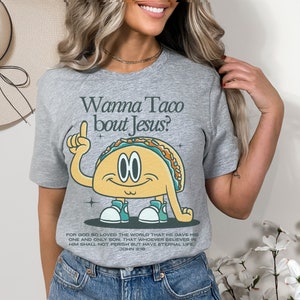 May include: Gray t-shirt with a cartoon taco wearing sneakers and pointing up. The text "Wanna Taco bout Jesus?" is above the taco. The text "FOR GOD SO LOVED THE WORLD THAT HE GAVE HIS ONE AND ONLY SON, THAT WHOEVER BELIEVES IN HIM SHALL NOT PERISH BUT HAVE ETERNAL LIFE. JOHN 3:16" is below the taco.