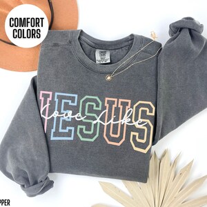 May include: A dark grey sweatshirt with the words "Jesus" in rainbow colours and "love like" in white script underneath. The sweatshirt is made by Comfort Colors.